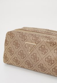 Brūna rakstaina kosmētikas soma ar teksturētu virsmu, zelta rāvējslēdzis un trīsstūrainais Guess logo accents sānos.
