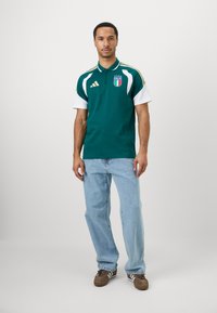 Mann som står iført en grønn og hvit Italia sportspolo, lyseblå jeans og brune joggesko med hvite striper på en ensfarget bakgrunn.