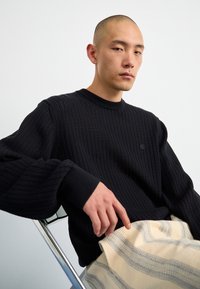 Černý žebrovaný pletený svetr s kulatým výstřihem a spadlými rameny, doplněný krémovými vzorovanými kalhotami. Jednoduchý design s texturovanou tkaninou.