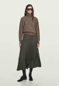 Brązowy sweter o fakturze z wysokim kołnierzem, zestawiony z ciemnozieloną plisowaną spódnicą. Modelka ma na sobie czarne skarpetki i buty, okulary przeciwsłoneczne oraz naszyjnik.