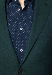 Zielona teksturowana marynarka nałożona na granatową koszulę z drobnymi białymi kropeczkami. Marynarka ma zapięcie na guziki oraz subtelne detale kołnierza.