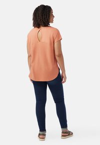 Top corto arancione chiaro a maniche corte con un'apertura a forma di chiave sulla schiena, abbinato a jeans skinny blu scuro e sandali marroni.
