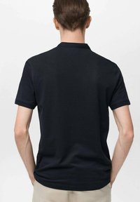 Černé polo tričko vyrobené z hladkého materiálu, má krátké rukávy a klasický límec. Pohled zezadu ukazuje přiléhavý design s minimalistickým stylem.