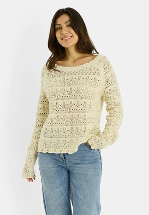 Joven sonriente, vestida con un suéter de crochet color crema y jeans azules, de pie con una mano en el bolsillo frente a un fondo liso.