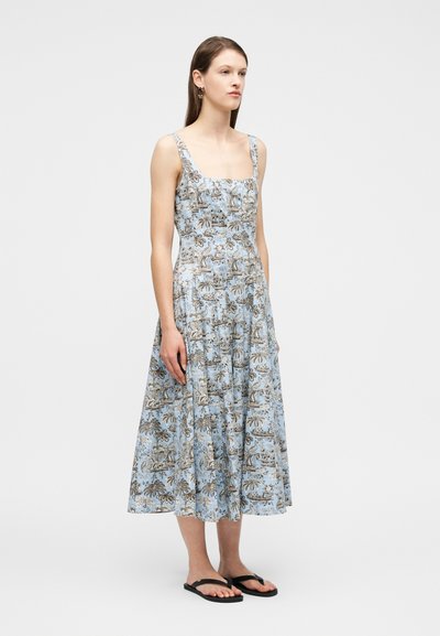 Νεαρή γυναίκα όρθια, φοράει αμάνικο, γαλάζιο midi φόρεμα με σχέδιο από φοινικόδεντρα και ζώα, και μαύρα σανδάλια με επίπεδο τακούνι.