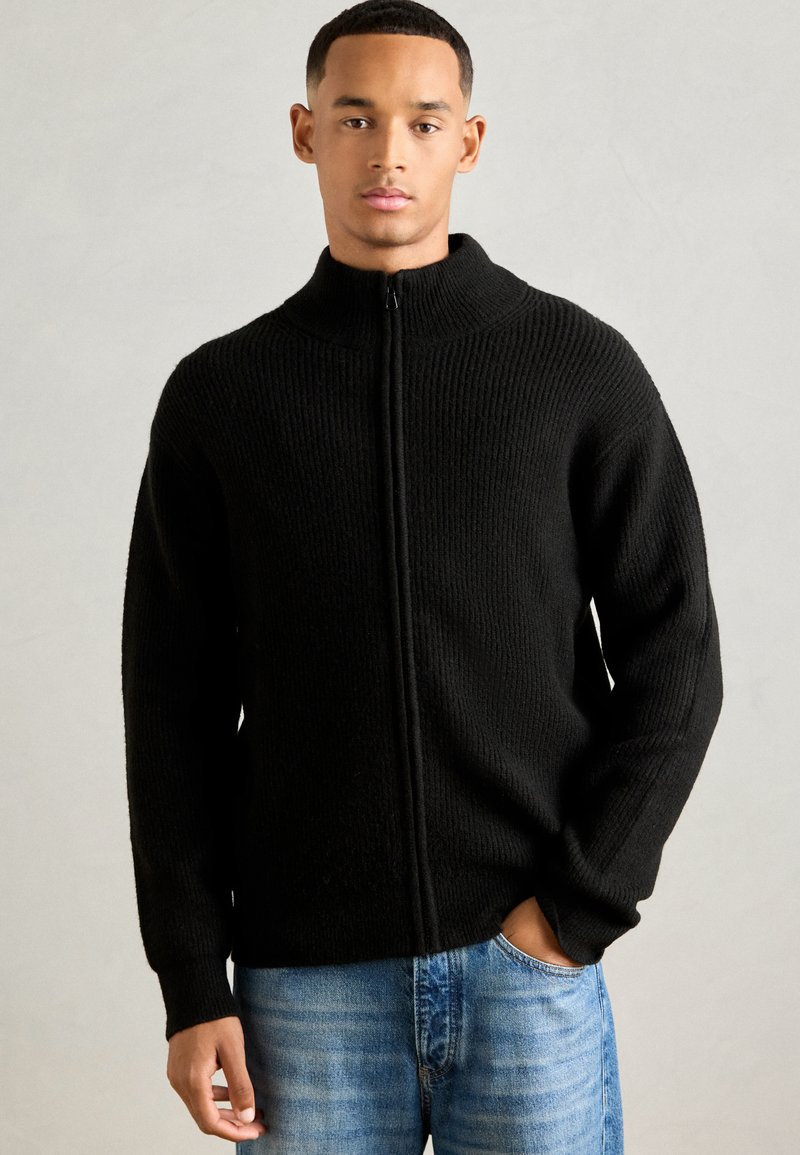 Jeune homme portant un pull en maille noir avec fermeture éclair et un jean bleu, se tenant contre un fond gris clair uni, une main dans sa poche.