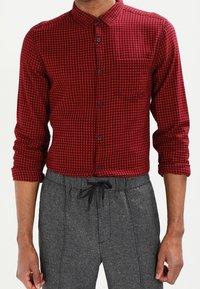 Camicia a quadri rossa e nera, design con bottoni, maniche lunghe arrotolate ai polsini, con una singola tasca sul petto, abbinata a pantaloni grigi con cordoncino.