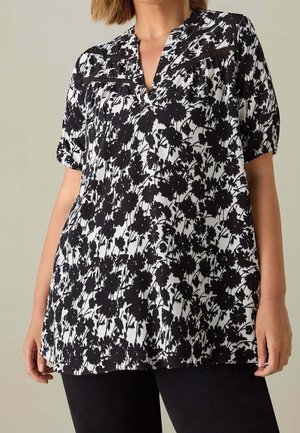 Frau trägt eine kurzärmelige Bluse mit schwarz-weißem Blumenmuster, V-Ausschnitt und Spitzenbesatz, kombiniert mit schwarzen Hosen.
