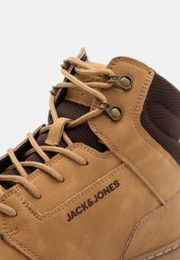 Bota de cuero marrón con acentos de malla en color marrón, que presenta cordones beige y ojales metálicos redondos. Logo "JACK & JONES" grabado en el costado.