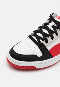 Schwarzer, weißer und roter Sneaker mit einer Obermaterial aus strukturiertem Leder, gelochtem Zehenbereich und schwarzen Schnürsenkeln, mit einem markanten roten Logoakzent.