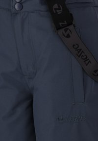 Gros plan sur un pantalon bleu foncé avec une taille à double bouton, une poche latérale et des bretelles noires de marque attachées.