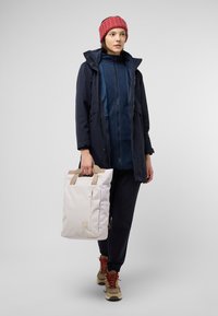Marineblauwe waterdichte parka over een blauwe fleece top, donkere broek, beige sneakers, met een crèmekleurige totebag met tan banden en een ritsvak.