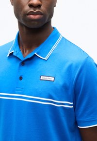 Άνδρας που φοράει φωτεινό μπλε πόλο μπλουζάκι με λευκές διπλές ρίγες στο στήθος και το γιακά, με λογότυπο Calvin Klein στην αριστερή πλευρά.