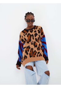 Maglione oversize color beige con stampa leopardata nera e marrone, con maniche a fantasia blu e rossa. Abbinato a jeans chiari strappati.