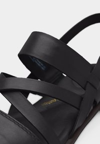 Κοντινό πλάνο μαύρου δερμάτινου σανδάλου με πολλαπλά φαρδιά και στενά λουριά που διασχίζουν το πόδι και τον αστράγαλο.