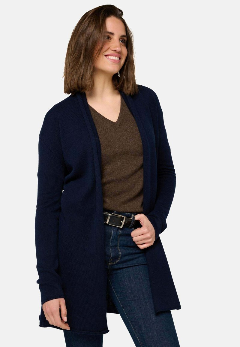 Cardigan long bleu marine en tissu tricoté, avec un devant ouvert, une coupe décontractée et des détails de poignets côtelés. Porté par-dessus un haut marron.