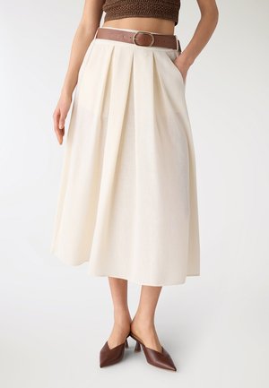 Kobieta ubrana w kremową plisowaną spódnicę midi z bocznymi kieszeniami, brązowy pasek, brązowe mulesy na obcasie z zaokrąglonym noskiem oraz krótki sweter.