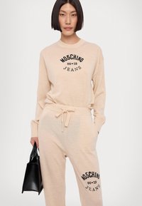 Conjunto de roupa de lazer bege, com mangas longas, decote redondo e calças com cordão. Logótipo preto "Moschino Jeans" no peito e na perna.