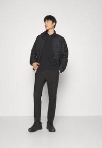 Chaqueta negra acolchada con acentos naranjas, combinada con una camiseta polo negra y pantalones negros. El modelo está de pie contra un fondo liso.