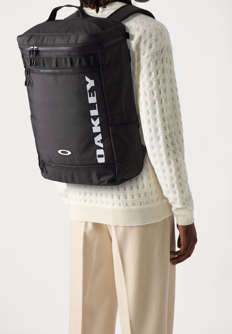 Personne portant un pull blanc texturé et un pantalon beige porte un grand sac à dos noir Oakley avec plusieurs compartiments zippés.