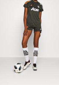 Zielona koszulka Manchester United z pomarańczowymi paskami, czarne spodenki, białe skarpety z czarnym wzorem oraz białe buty piłkarskie. Obok piłka nożna.