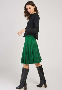 Pull noir à manches longues associé à une jupe plissée verte et texturée. Le mannequin porte des bottes noires à hauteur de genou avec un talon carré.