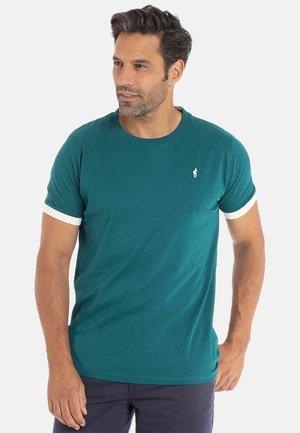 Homme aux cheveux courts et foncés portant un t-shirt court teal avec des bords de manches blancs et un pantalon sombre, regardant sur le côté.