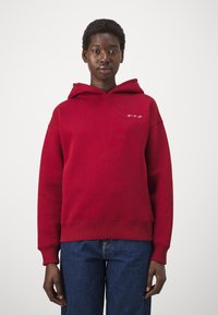 Roter Kapuzenpullover mit einem weißen Logo auf der Brust. Hergestellt aus Baumwolle, hat er eine lässige Passform sowie gerippte Bündchen und Saum.