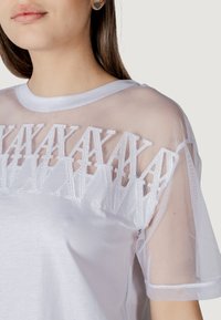 Camiseta blanca con mangas de malla transparente, que presenta patrones geométricos bordados en el pecho. Textura de tela suave, cuello redondo.