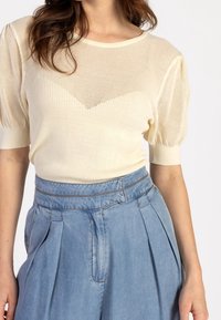 Světle béžový síťovaný svetr s krátkými, vypouklými rukávy; strukturovaná žebrová vazba v vzoru šipky. Spárováno s modrými džíny s vysokým pasem.