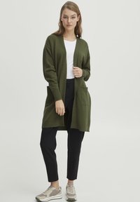 Long cardigan vert olive avec une fermeture ouverte, des poignets côtelés et deux poches avant, associé à un pantalon noir et des baskets beiges.
