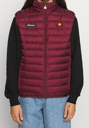 Persoon draagt een bordeauxrood gewatteerd vest met ritssluiting, zwarte mouwen en lichtblauwe spijkerbroek, met ellesse-logo badges op de borst.