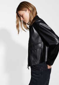 Giacca in pelle nera con chiusura a zip e tasca frontale. Texture liscia, silhouette leggermente oversize, presentata su uno sfondo bianco.