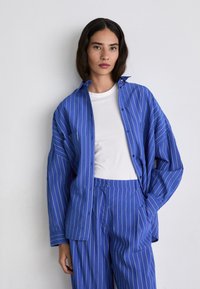Camicia oversize a righe blu con chiusura a bottoni, abbinata a pantaloni a righe blu coordinati e una maglietta bianca a girocollo.