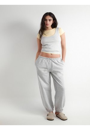 Młoda kobieta z długimi ciemnymi włosami, ubrana w żółtą koszulkę pod szarym topem, szare spodnie dresowe i beżowe buty wsuwane.