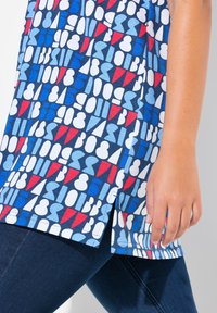 Haut bleu à motifs avec des formes abstraites en rouge, blanc et bleu clair, avec une fente sur le côté et des manches courtes. Associe à un jean foncé.