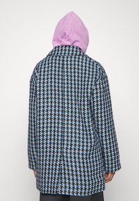 Upletený kabát s vzorem houndstooth v černé, modré a bílé barvě. Obsahuje růžovou kapuci a volný střih s dlouhými rukávy.