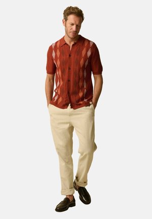Homme portant une chemise à manches courtes couleur rouille avec un motif en losange, un pantalon beige retroussé et des mocassins marron foncé, les mains dans les poches.
