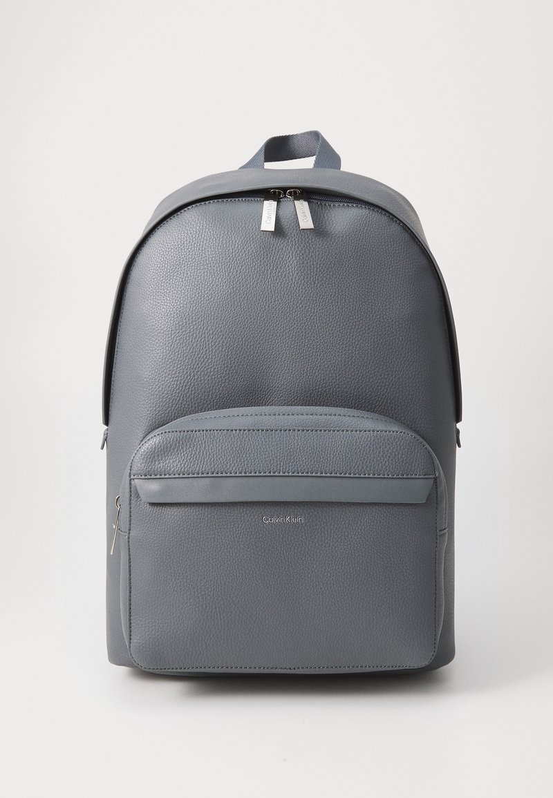 Sac à dos en cuir texturé gris Calvin Klein avec poche avant zippée, double fermeture éclair sur le compartiment principal et poignée de transport supérieure.