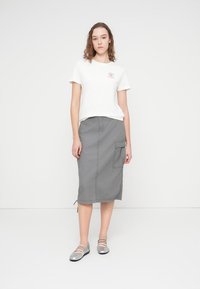 Γκρι midi φούστα με πλαϊνή τσέπη, συνδυασμένη με λευκό T-shirt που φέρει μικρό τυπωμένο λογότυπο. Το μοντέλο φορά ασημένια μπαλαρίνες.