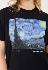 Camiseta preta em algodão com uma impressão de uma pintura famosa, predominantemente azul com padrões ondulantes, uma torre proeminente e o texto "Noite Estrelada, Vincent van Gogh."
