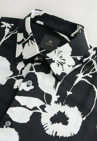 Schwarzes Hemd mit einem floralen weißen Muster, ausgestattet mit einem klassischen Kragen, Knopfverschluss und glatter Stoffoptik. Markenetikett sichtbar.