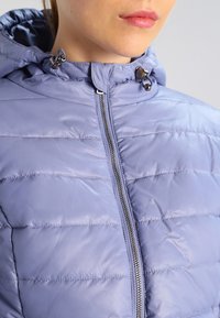 Ljuslila pufferjacka med huva, som har horisontell sömnad, en silverdragkedja och justerbara snoddar vid kragen.