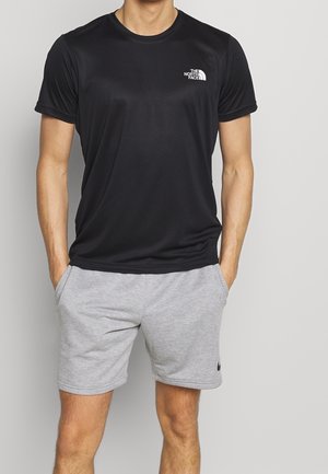 Mężczyzna w czarnej koszulce The North Face i jasnoszarych spodenkach Nike, z rękami w kieszeniach, na jednolitym tle.