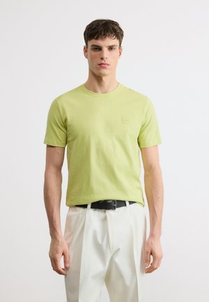 Jeune homme portant un t-shirt à manches courtes vert clair et un pantalon blanc plissé avec une ceinture noire, debout devant un fond uni.