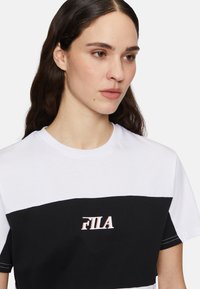 Vit och svart kortärmad t-shirt med ett horisontellt färgblockdesign och FILA-logotyp i röd kontur, med rund halsringning och mjuk textur.