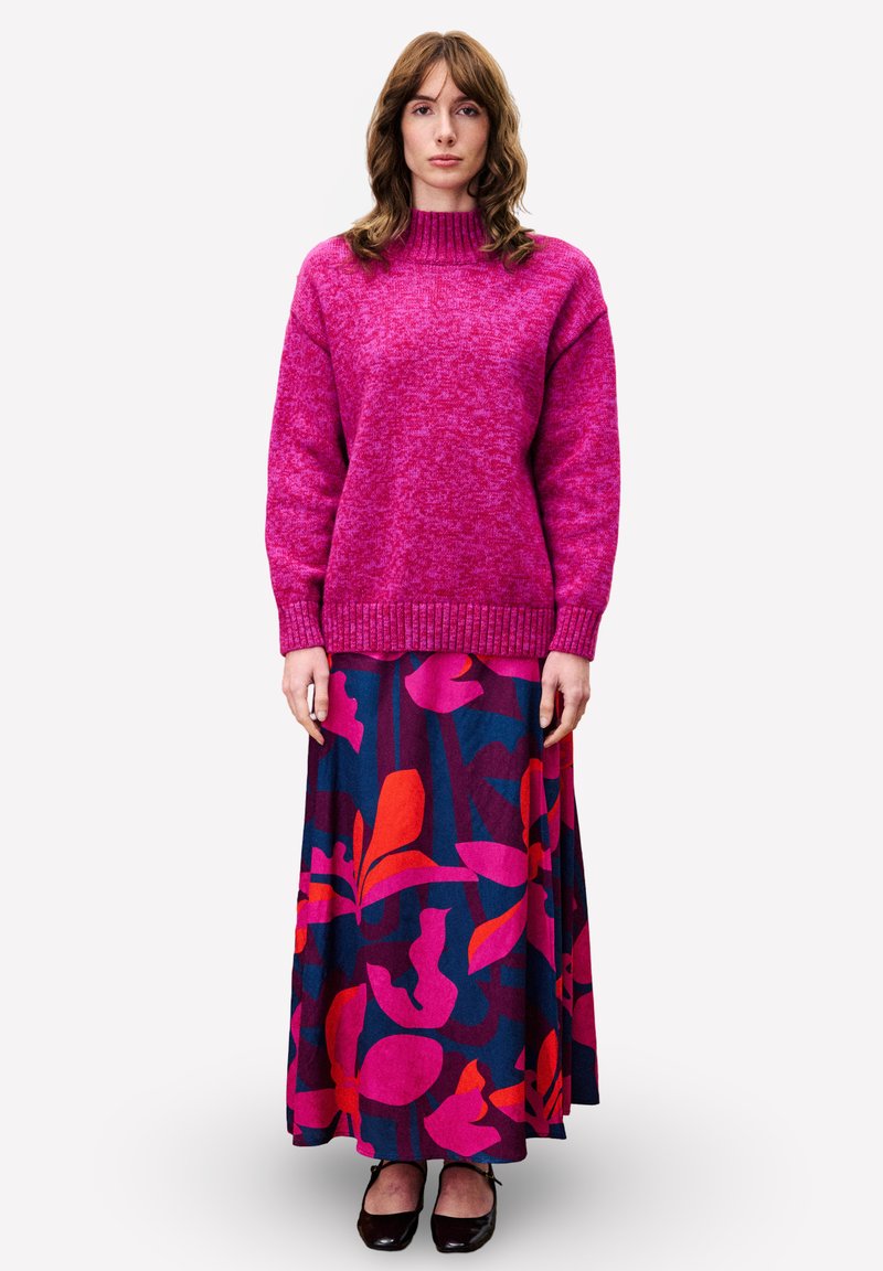 Vrouw in een felroze gebreide trui en een lange marineblauwe rok met grote roze en rode bloemenpatronen, staand met ontspannen armen.