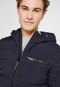 Marineblaue Steppjacke mit Kapuze, gestepptem Design, Frontreißverschluss und Brusttasche; hergestellt aus leichtem, strukturiertem Material.