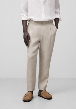 Person trägt beige maßgeschneiderte Hose, weißes langärmeliges Hemd und braune Slipper, steht vor einem einfarbigen hellgrauen Hintergrund.