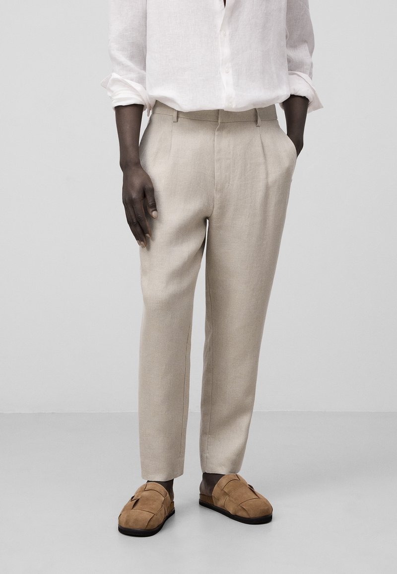 Personne portant un pantalon beige sur mesure, une chemise blanche à manches longues et des chaussures marron sans lacets, debout devant un fond uni gris clair.